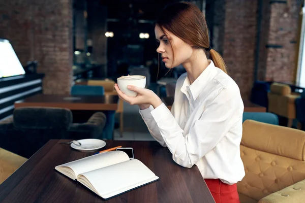 Mulher bonita com uma xícara na mão senta-se a uma mesa em um café e reservar um restaurante — Fotografia de Stock