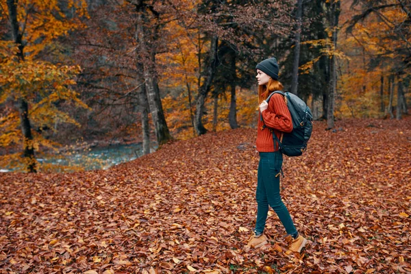 快乐的女旅行家背着背包在大自然的秋日公园里散步，在河边的风景旁边，高大的树毛衫 — 图库照片