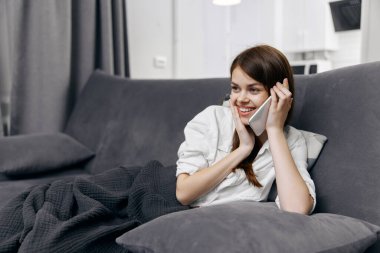Kanepede oturan bir kadın. Gri perdeler. Cep telefonu iletişim ekoseli.