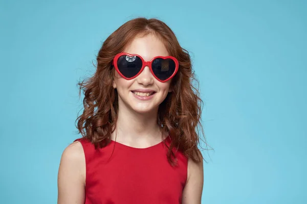 Malá dívka v slunečních brýlích v podobě srdcí červené šaty kudrnaté vlasy modré pozadí — Stock fotografie