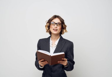 Elinde bir kitapla ceketli bir kadın yüzünde kıvırcık saçlı, açık renk bir gözlük takıyor.