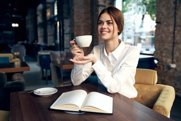 손에 컵을 든 행복 한 여인 이 식당의 의자에 앉아 있고 뒤에는 내부가 있다 — 스톡 사진