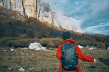 Kırmızı ceketli, sırt çantalı ve sıcak şapkalı bir kadın sonbaharda dağlarda çimlerin üzerinde yürüyor. 