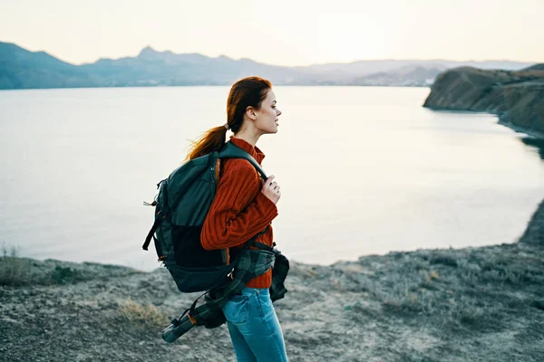 Glückliche Frau in rotem Pullover mit Rucksack auf dem Rücken, die sich in der Natur in der Nähe des Meeres und der Berge in der Ferne ausruht — Stockfoto