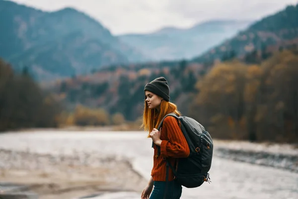 Mulher viajante com uma mochila e em um chapéu perto do rio nas montanhas no outono — Fotografia de Stock