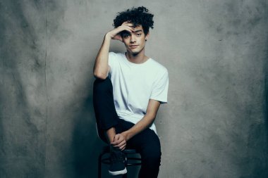 Beyaz tişörtlü adam sandalyeye oturur. Kıvırcık saç stili.