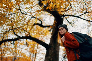 Kırmızı kazaklı bir sırt çantasıyla mutlu yolculuklar ve ağaçların yanındaki parkta kot pantolonlar.