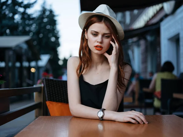 街上一家咖啡馆里，一个头戴白色帽子，身穿黑色连衣裙的女人坐在桌子旁 — 图库照片