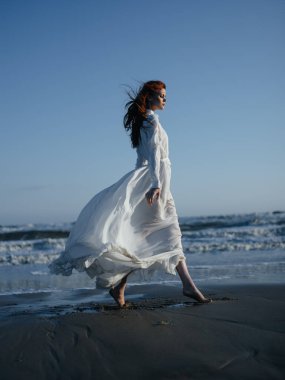 Beyaz elbiseli bir kadın okyanusun kıyısındaki ıslak kumda yürüyor.