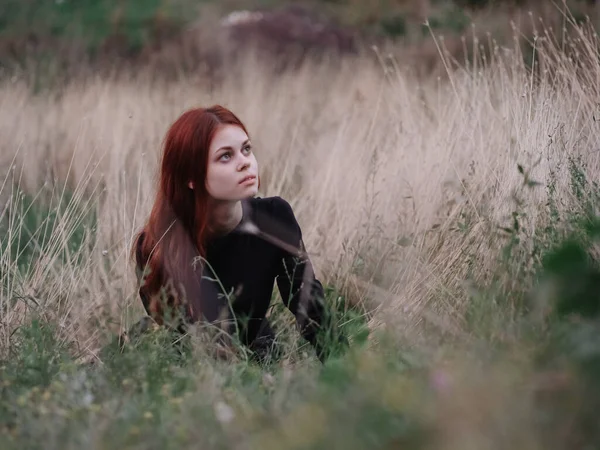 Vörös hajú nő fekete ruhában fekszik az erdőben száraz füvön a természetben — Stock Fotó