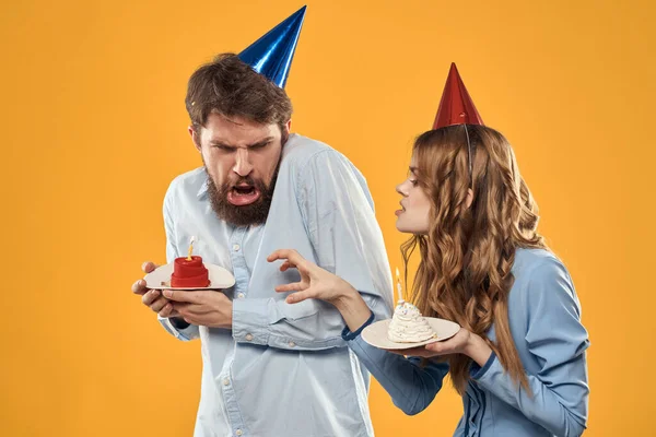 Sarı arka planda pastası olan şapkalı kadın ve erkek doğum günü partisi. — Stok fotoğraf