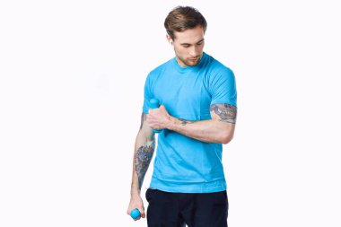 Kolunda mavi tişörtlü ve kaslı vücut geliştirme formundaki dövmeli bir adam.