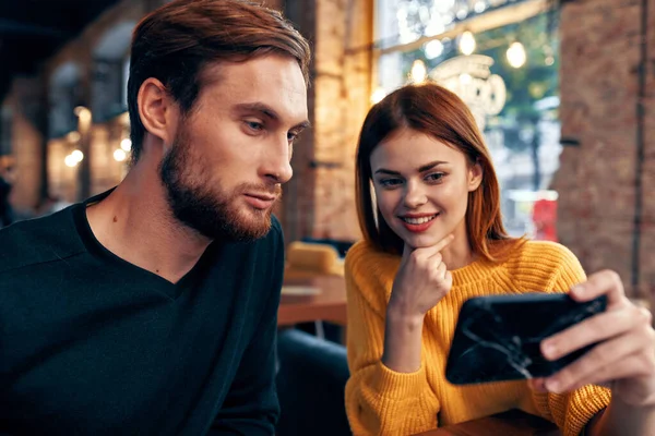 Una mujer en un suéter con un teléfono móvil y un tipo con barba están sentados en un restaurante —  Fotos de Stock