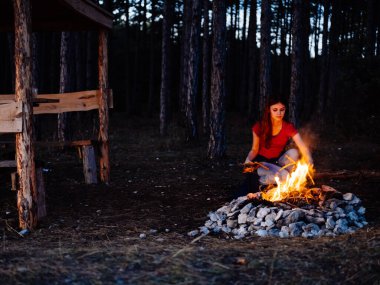 Woman near a campfire outdoors in the evening in a pine forest clipart