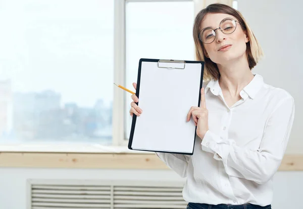 Eine Frau hält einen Ordner mit einem weißen Blatt Papier und einem Fenster im Hintergrund — Stockfoto