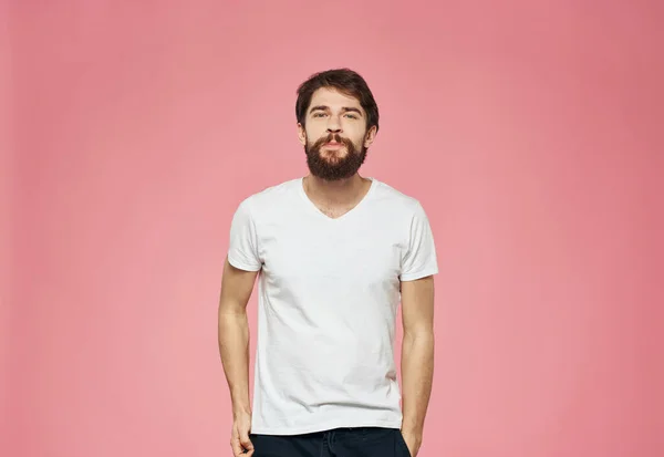 Um cara com barba em um fundo rosa, uma camiseta branca e calças escuras — Fotografia de Stock