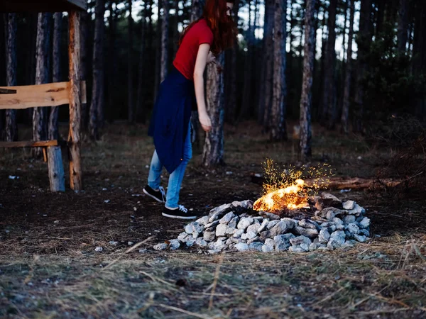 Una mujer romántica en jeans y una camiseta se sienta cerca de una fogata en la naturaleza —  Fotos de Stock