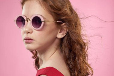 Kıvırcık saçlı, siyah gözlüklü, eğlenceli kırmızı elbiseli, pembe arka planlı bir kız.