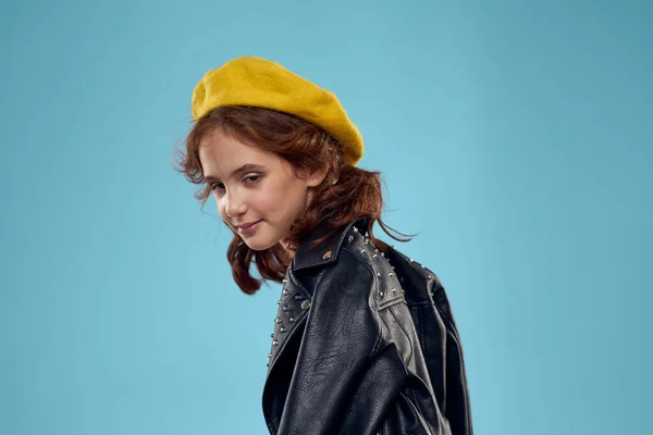Modieus meisje in een zwart leren jasje op een blauwe achtergrond en een gele muts op haar hoofd — Stockfoto
