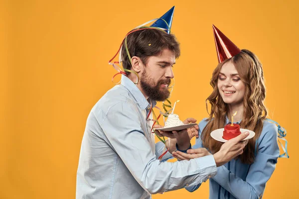 Homem e mulher em uma festa em bonés e com bolo de ouropel divertido fundo amarelo — Fotografia de Stock