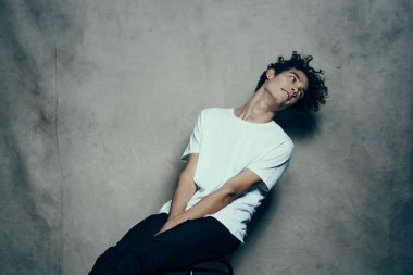 Kıvırcık saçlı bir genç, kumaş arka planda bir sandalyede oturur.