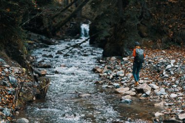 Sırt çantalı bir kadın turist nehir kıyısında yürüdü, bir dere dolusu su ve uzun ağaçlar.