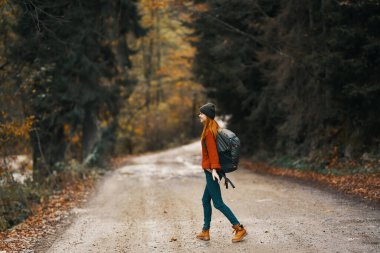 Kot ceketli bir kadın sonbahar ormanında yol boyunca sırt çantalı yürüyor.