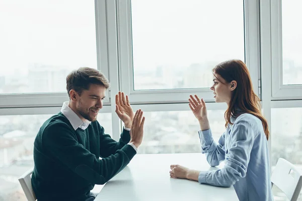 Ein Mann im Pullover und eine Frau im Hemd gestikulieren mit den Händen und sitzen an einem Tisch am Fenster — Stockfoto