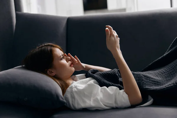 Ładna kobieta leży na kanapie pokryta kocem i patrzy na telefon komórkowy — Zdjęcie stockowe