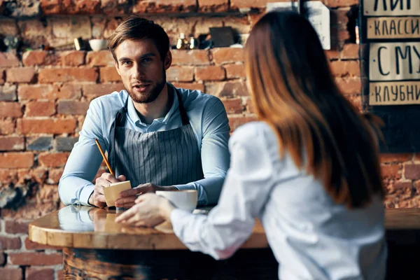 회색 앞치마를 입은 남성 웨이터 가 카페에 있는 테이블에서 여성 손님을 위해 주문을 받고 커피를 마시는 모습 — 스톡 사진