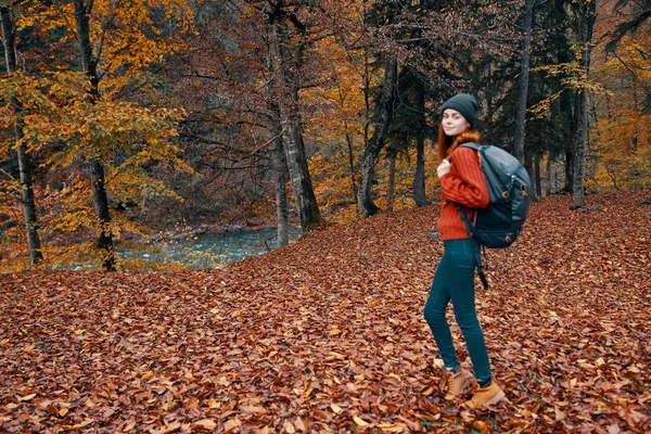 Jovem feliz com uma mochila em botas de jeans e uma camisola estão andando na floresta de outono perto das árvores altas — Fotografia de Stock