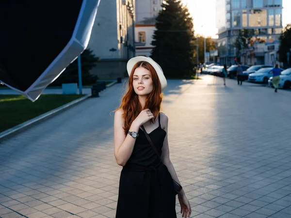 Modelo de mulher ruiva na rua na cidade em um vestido preto e em um chapéu branco — Fotografia de Stock