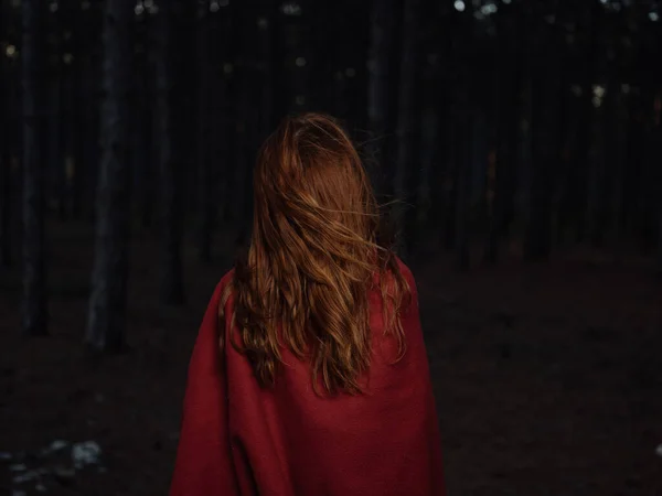Una mujer viaja en el bosque por la noche con una tela escocesa roja sobre sus hombros, vista hacia atrás —  Fotos de Stock