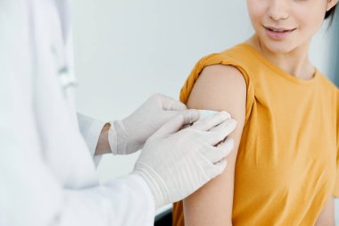 Koruyucu eldivenli doktor sarı tişörtlü bir kadının omzunu bantlıyor.