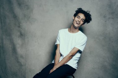 Kıvırcık saçlı bir genç, kumaş arka planda bir sandalyede oturur.