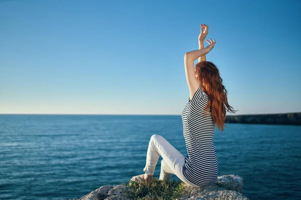 woman with hands raised up sits on high stones near the sea in summer