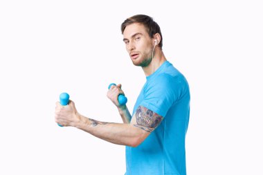 Mavi tişörtlü spor adamı elinde dambıllar, spor salonu manzarası.