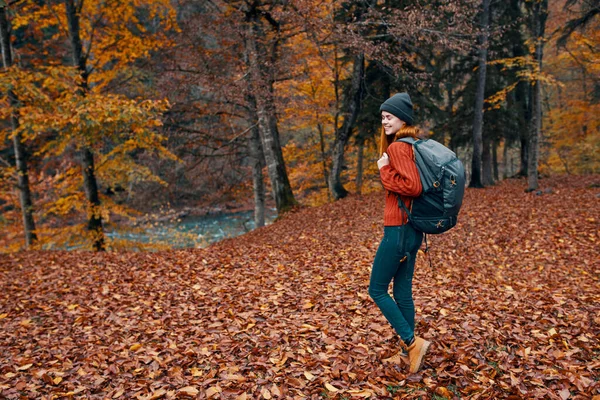 Glückliche junge Frau mit Rucksack in Jeansstiefeln und Pullover spazieren im herbstlichen Wald in der Nähe der hohen Bäume — Stockfoto