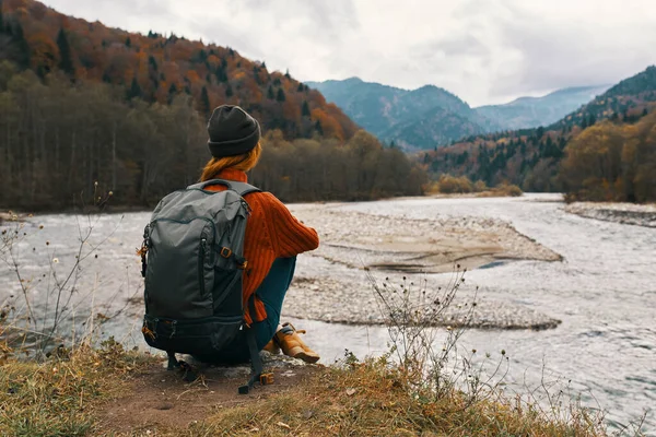 Mujer en las montañas en otoño con una mochila se sienta en la orilla del río y mira a las altas montañas —  Fotos de Stock
