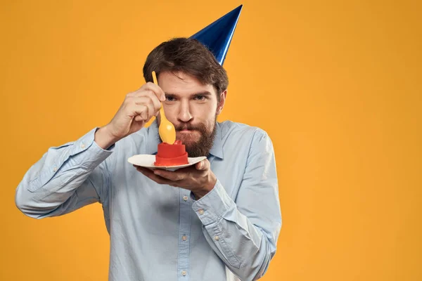 Un hombre con un pastel de cumpleaños en sus manos y con una gorra sobre un fondo amarillo en su cumpleaños — Foto de Stock