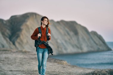 Sırtında sırt çantası olan bir kadın turist dağlardaki deniz kenarında yürüyor.