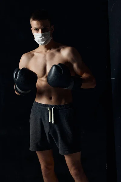 Sportovní muž v lékařské masce a v boxerských rukavicích na černém pozadí kraťasy fitness model — Stock fotografie