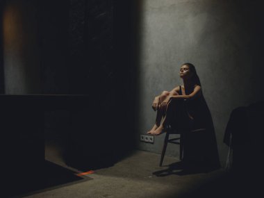 Karanlık bir odada, yalnız üzgün bir kadın oturuyor dirsekleri duvarda ve elleriyle bacaklarına sarılıyor.