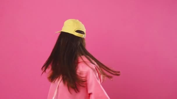 Happy woman in yellow cap and pink sweatshirt jumping and laughing in front of the camera in the studio on a pink background — Stock Video