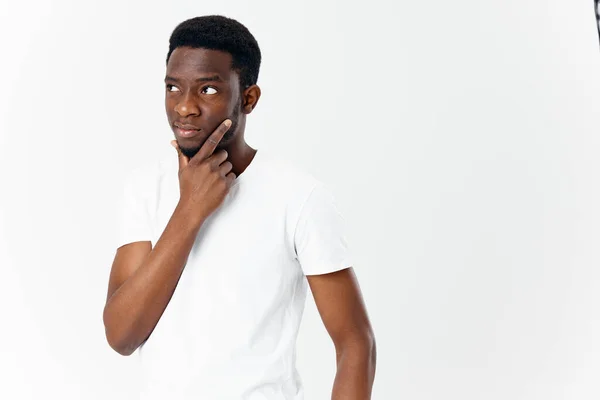 Verbijsterd afrikaanse man kijkt naar de zijkant raakt zijn gezicht met zijn hand op een lichte achtergrond — Stockfoto