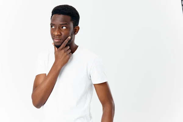 Desconcertado hombre africano mirando a un lado toca su cara con su mano sobre un fondo claro — Foto de Stock