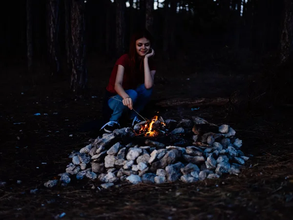 Una mujer romántica en jeans y una camiseta se sienta cerca de una fogata en la naturaleza —  Fotos de Stock