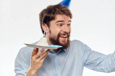 Elinde pasta tabağı ve açık arka planda mavi gömlekli bir adam doğum günü partisi şapkası takıyor.