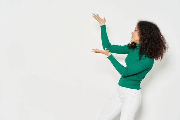Krullende mulatto vrouw in een groene trui gebaren met haar handen op een lichte achtergrond Copy Space — Stockfoto