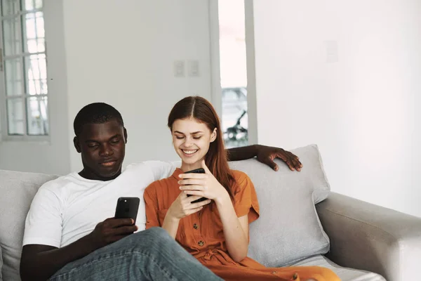 Femme en robe de soleil et un homme africain avec des téléphones mobiles sont assis sur le canapé bavarder intérieur de la famille — Photo